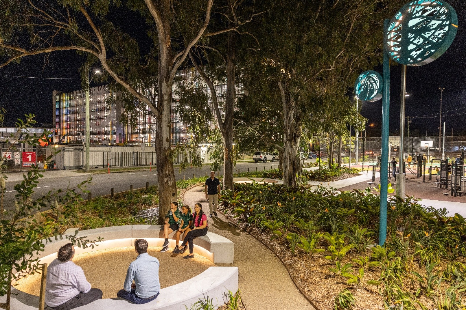 People seated and walking along a landscaped, well‑lit pedestrian path at night, featuring curved concrete seating, trees, garden beds, and illuminated public art, with buildings and street lighting visible in the background