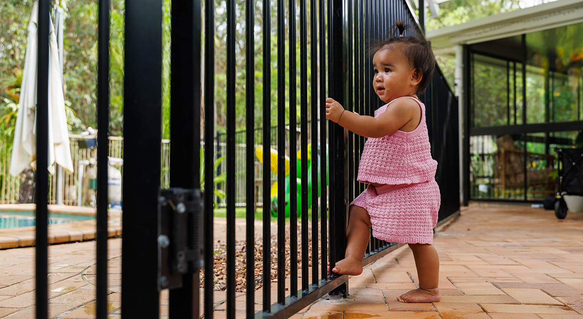 Toddler leaning against a rail, the pool is behind it and unreachable to her, keeping her safe