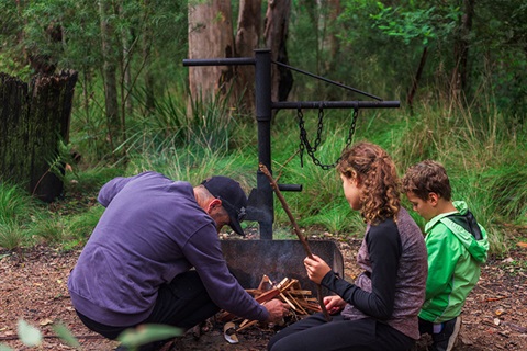 Man and two teenage children kneel around fire pit as man stacks kindling