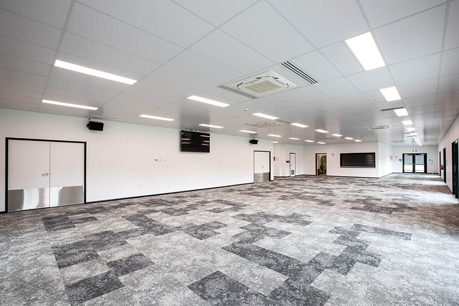 Large room with multiple doorways and grey patterned carpet, flat screen tv and speakers are mounted on one wall.