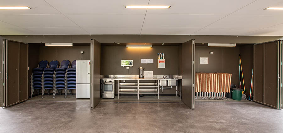 Open cupboards showing rows of blue stacked chairs to the left, a kitchen area with a small fridge, bench and sink and to the right, fold up tables and cleaning equipment.