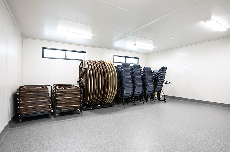 Room with two rectangle shaped windows with rows of blue chairs and brown tables stacked against one wall.