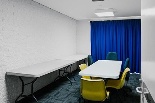 White bick room. Carpeted with a blue curtain and some tables and chairs.
