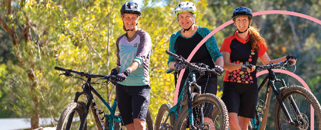 Three women smile while standing beside their bikes