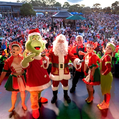 Santa Clause and the grinch with a group of elves standing on a stage at a carols event