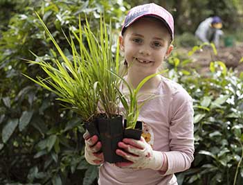 Girl wearing pink jumper and a cap holds up large plant and smiles