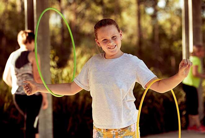 A young girl smiling at the camera with a hula hoop around each arm.