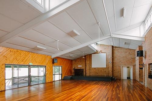 large hall with brick walls and wooden floors. small stage at front of room.