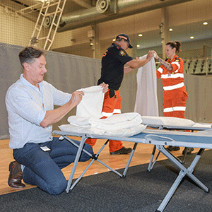 Man folds sheets with a camp stretcher while two volunteers also help set up an evacuation centre.