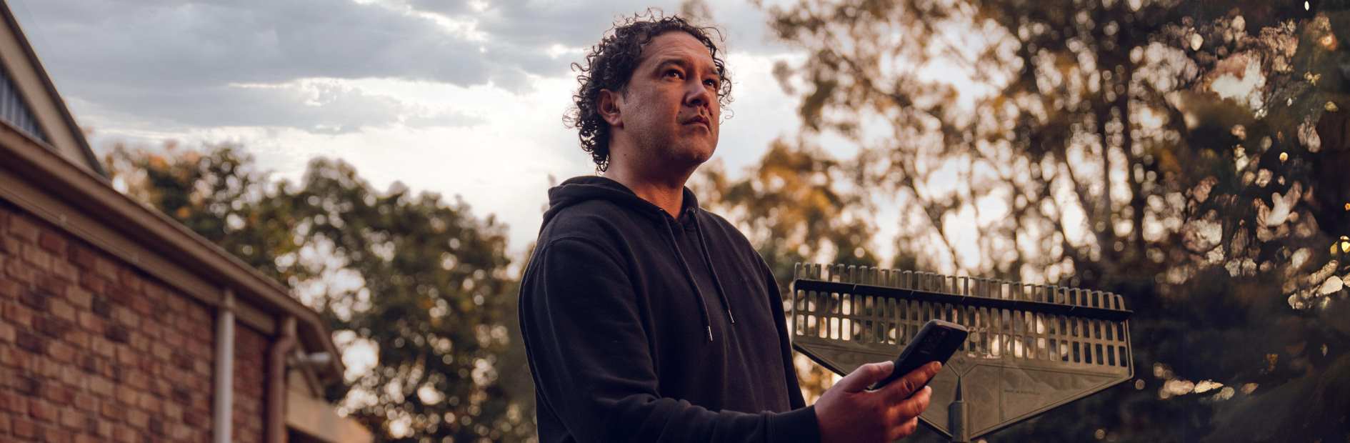  Man stands outside house holding a rake and a phone.