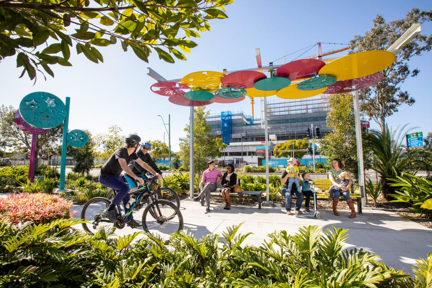 Two people riding their bikes on the new healthy street paths, behind them people are seen sitting below a colourful shade structure. In the background additional disk structures are visible among the greenery.