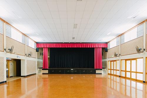 Community hall with wooden look flooring and brick walls. large stage at front of the room with red curtains