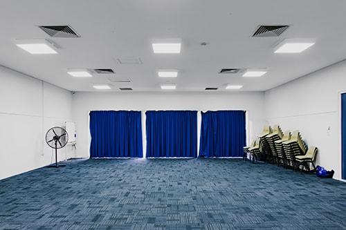 White room with blue carpet and curtains, chairs piled in far corner.