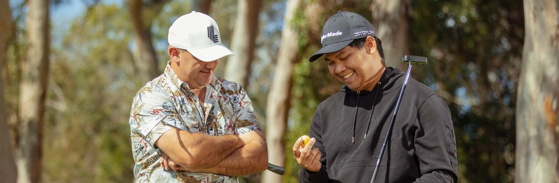 Two smiling men dressed in polo shirts and caps hold golf clubs and inspect a yellow golf ball