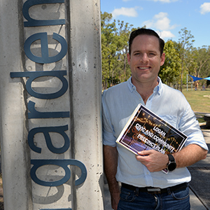 An image of Cr Jon Raven, holding the draft plan and leaning against the entrance sign to Logan Gardens