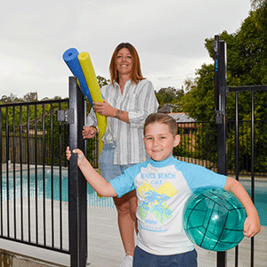 mum holds pool noodles and stands at gate to pool while son holds ball