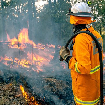 Person dressed in orange firecoat and helmet stands in front of flames holding a hose