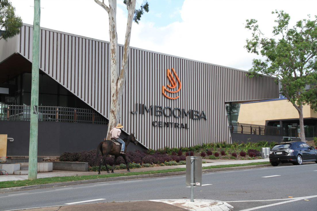 Picture of the Jimboomba Central project showing part of the road and the outside of the building with a man riding a horse on the footpath