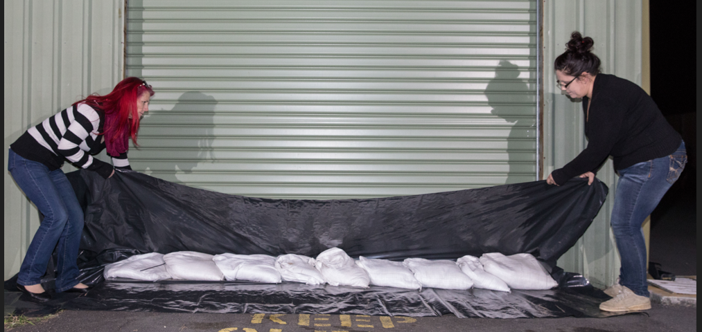 Two people who have laid black plastic along the door opening of a shed and are putting sandbags on the plastic to avoid water entering the shed.