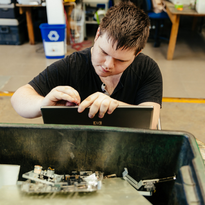 A boy pulling apart a laptop with a bucket in front of him where he is putting the parts for recycling