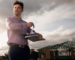 Man with clipboard stands in front of half finished house with wooden frame work exposed
