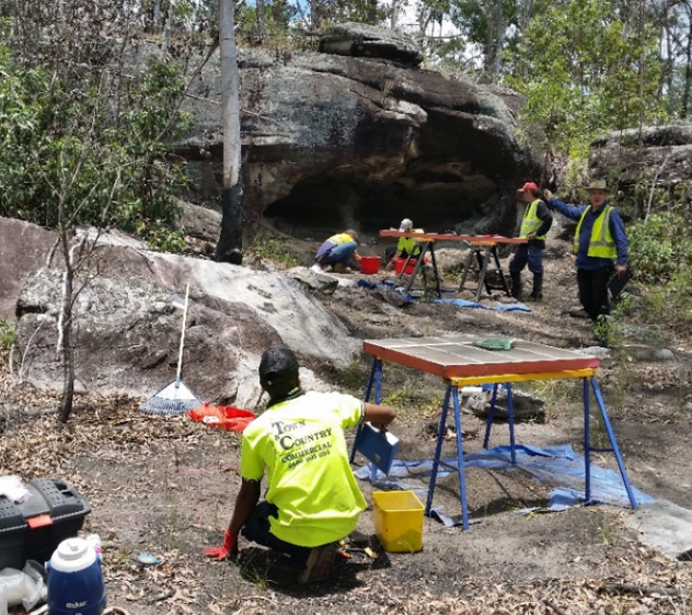 In the foreground a Jabree team member is kneeling beside a workstation excavating an area. In the background four Jabree team members are conducting excavations directly in front of the Kitchen Shelter.