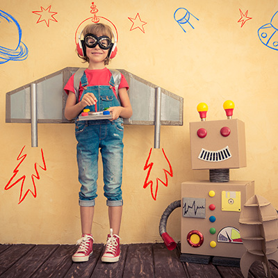 Child dressed up with cardboard crafted wings next to a cardboard crafted rocket and decorated cardboard box robot