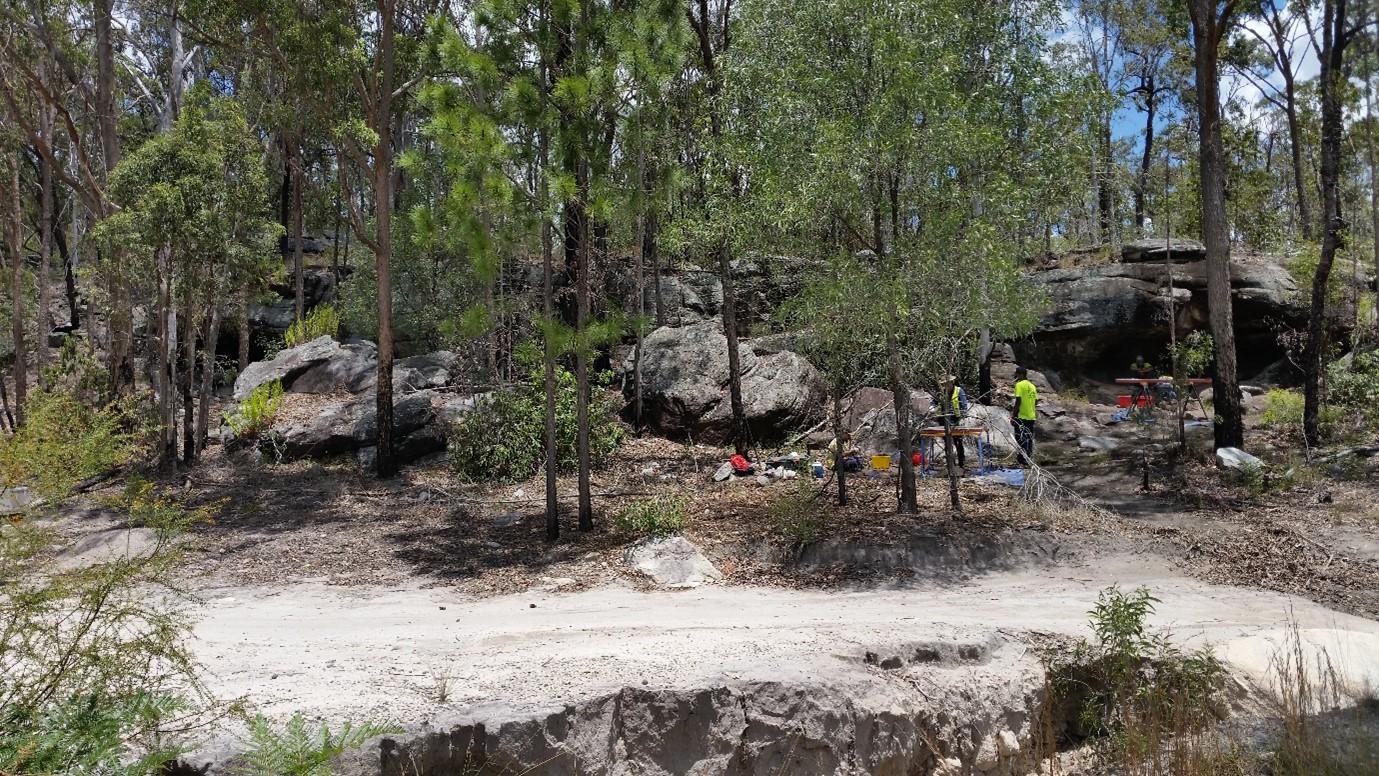 The Tool Making Shelter located on the left semi-obscured by trees is the smaller of the two rock shelters. It was excavated in 2011. The Kitchen Shelter located on the right is the larger rock shelter. It is surrounded by trees. Two people are working close to the entrance of the Kitchen Shelter. They are investigating the subsurface (2016).